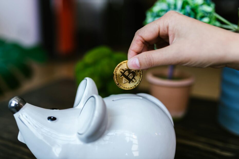 A hand placing a Bitcoin coin into a white piggy bank symbolizing savings.